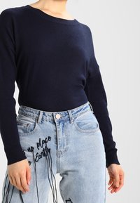 Maglione a maglia blu navy con scollatura ampia abbinato a jeans azzurri chiari con testo ricamato in nero e vestibilità comoda.