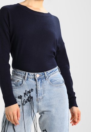 Marineblauer Strickpullover mit weitem Halsausschnitt kombiniert mit hellblauen Jeans, die schwarzen Sticktext haben und einen lässigen Schnitt bieten.