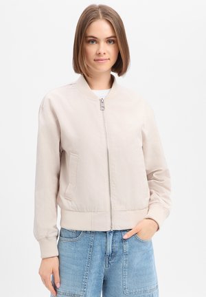 Młoda kobieta z włosami do ramion, ubrana w jasnobeżową kurtkę typu bomber zapinaną na zamek oraz niebieskie dżinsy, stoi z jedną ręką w kieszeni.