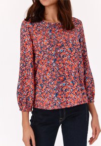 Rød blomstret bluse med lange, puffede ærmer. Har en rund halsudskæring og et miks af små blå, hvide og orange blomster på en lys baggrund.