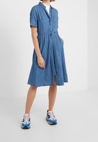 Denimová košilová šaty s krátkým rukávem, opaskem v pase, přední kapsou a sukní do délky kolen. Nosené s barevnými sportovními botami.