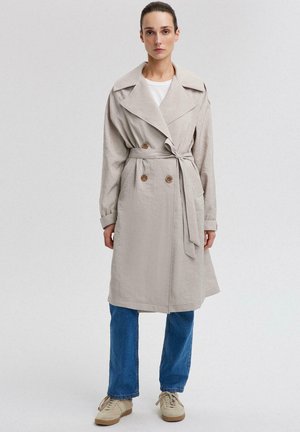 Beige double-breasted trench coat with large lapels, belted waist, two front pockets, and structured fabric; paired with blue jeans and sneakers.