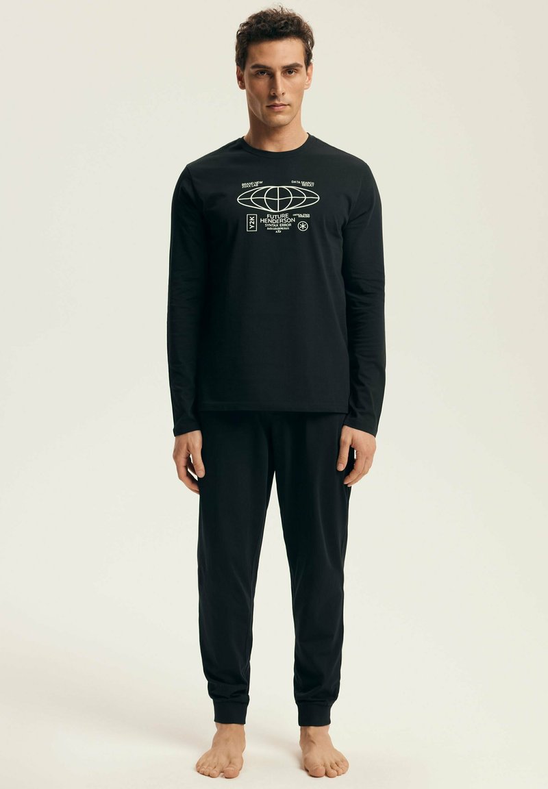 Man stands barefoot wearing black long-sleeve shirt with white tech graphic and matching black jogger pants against a plain light background.