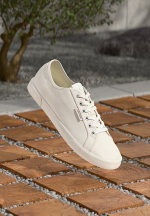 Fehér vászon sneaker kerek orral, lapos fehér gumitalppal és ezüst fűzőlyukakkal. Oldalán egy kis logótábla található.