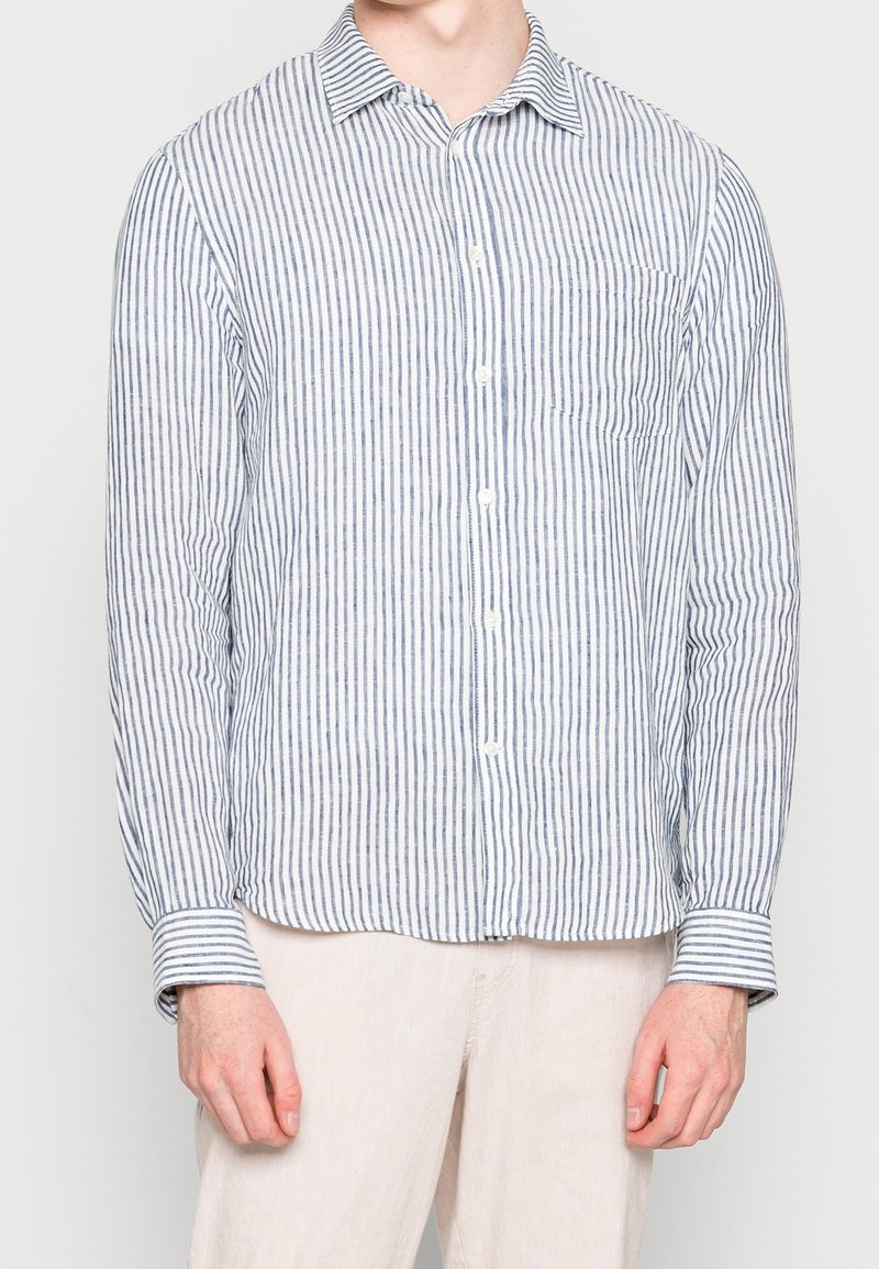 Homme portant une chemise à manches longues boutonnée avec des rayures verticales bleues et blanches et un pantalon beige clair, debout devant un fond uni.