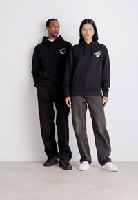 Sudaderas negras con bolsillos delanteros y cordones, usadas por dos modelos, combinadas con pantalones negros y grises, sobre un fondo liso.