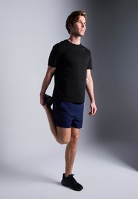 Zwarte t-shirt met korte mouwen en marineblauwe shorts gedragen door een man die één been strekt, met sportieve schoenen en een lichte huidtoon.