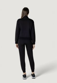 Tuta sportiva nera con giacca aderente e pantaloni affusolati. Il materiale appare liscio. Presenta polsini e orlo a coste, abbinata a sneakers nere.