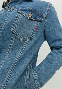 Blaue Jeansjacke mit Brustklappentasche, die einen kupferfarbenen Knopf und ein kleines rotes Punktdetail am Rand der Tasche aufweist.