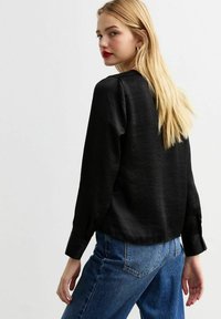 Blusa nera a maniche lunghe con finitura texturizzata, scollatura rotonda e polsini con bottoni, abbinata a jeans blu a vita alta.