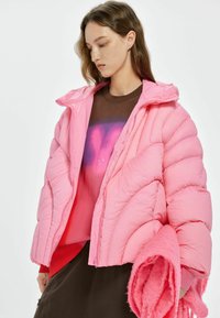 Roze, pufferjack met een cropped ontwerp en een gewatteerd patroon. Draag het over een bruine sweatshirt met een gradient kleurverloop, gecombineerd met een fluffy roze accessoire.