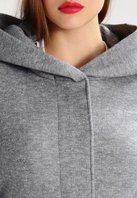 Vrouw draagt een grijze kleding met capuchon, met zichtbare rode lippenstift, die nek en onderkant van het gezicht toont zonder ogen of haar te onthullen.