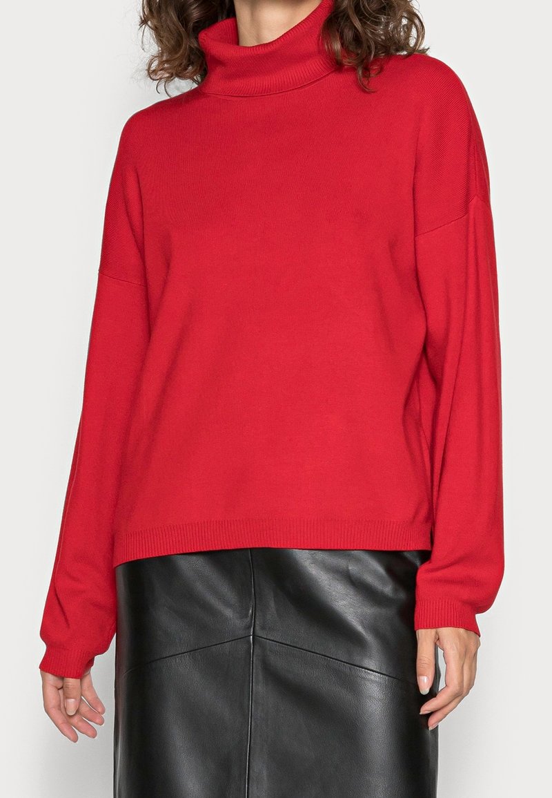Czerwony sweter typu golf w miękkim dzianinowym materiale, z opadającymi ramionami i ściągaczami przy rękawach, zestawiony z czarną skórzaną spódnicą.