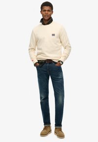 Beige sweatshirt met logo, gedragen over een groen geruit overhemd. Blauwe jeans met versleten plekken, gecombineerd met beige veterschoenen. Casual outfit.