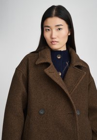 Καφέ παλτό σε σχέδιο ψαροκόκκαλο με φαρδύ γιακά, διπλό κούμπωμα και σκούρα κουμπιά. Φοριέται πάνω από ένα ναυτικά μπλε πουλόβερ με υφή.