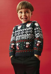 Maglione di Natale lavorato a maglia con sfondo blu navy, decorato con facce di Babbo Natale, fiocchi di neve e motivi festivi in rosso, verde e bianco. Pantaloni neri.