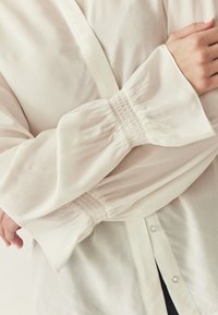 Blouse blanche à manches longues froncées avec smocks aux poignets. Le tissu semble doux et léger, avec une fermeture à boutons.