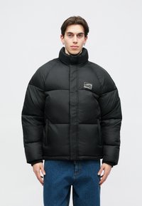 Μαύρο μπουφάν με ψηλό γιακά, με λευκό λογότυπο στο στήθος, με καπιτονέ υφή και δύο μπροστινές τσέπες.