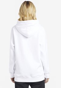 Persona bionda con capelli lunghi fino alle spalle, che indossa una felpa bianca semplice e pantaloni neri, voltata di spalle su uno sfondo bianco.