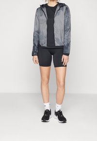 Giacca grigia leggera con cappuccio e finitura liscia, abbinata a una t-shirt nera e pantaloncini neri. Scarpe sportive nere con dettagli bianchi.