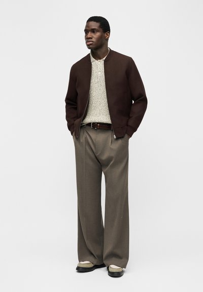 Νέος άνδρας που φοράει καφέ ζακέτα με φερμουάρ, ανοιχτόχρωμο πλεκτό πουκάμισο, παντελόνι ταμπά με φαρδιά πόδια και δίχρωμα παπούτσια μπεζ και μαύρα, στέκεται με τα χέρια στις τσέπες.