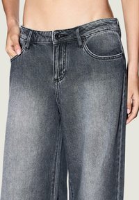 Jeans de mezclilla gris de pierna ancha con un lavado descolorido, bolsillos delanteros y costuras en contraste. La cintura cuenta con un botón y presillas para el cinturón.
