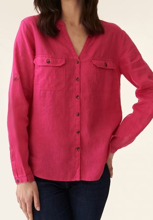 Kobieta w jasnoróżowej, długorękawowej koszuli na guziki z dwoma kieszeniami na piersi i ciemnych spodniach, stoi z jedną ręką na biodrze.