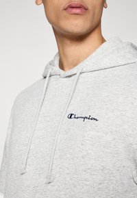 Γκρι φούτερ με απαλό υλικό, με κουκούλα με κορδόνι και κεντημένο λογότυπο "Champion" σε ναυτικό μπλε. Κοντινό πλάν ο στον γιακά και την περιοχή του στήθους.
