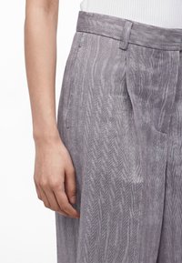 Το αριστερό χέρι ακουμπάει στο πλάι, φοράει γκρι παντελόνια με ψηλή μέση και σχέδιο ψαροκόκαλο και λευκό ριμπ τοπ που είναι μπλουζωμένο μέσα.