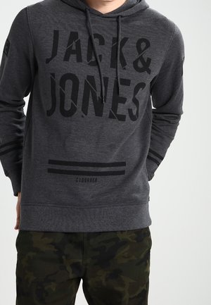 Hombre con sudadera con capucha Jack & Jones gris oscuro con texto negro y pantalones de camuflaje, de pie con las manos detrás de la espalda.