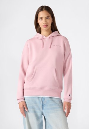 Junge Frau trägt einen hellrosa Champion Hoodie mit Fronttasche und hellblauen Jeans, steht vor einem schlichten weißen Hintergrund.