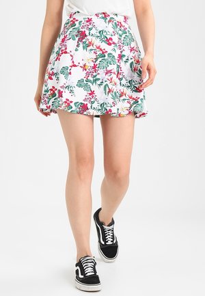 Femme portant une mini-jupe blanche à fleurs rouges, vertes et jaunes, associée à des baskets noires et blanches, debout devant un fond uni.