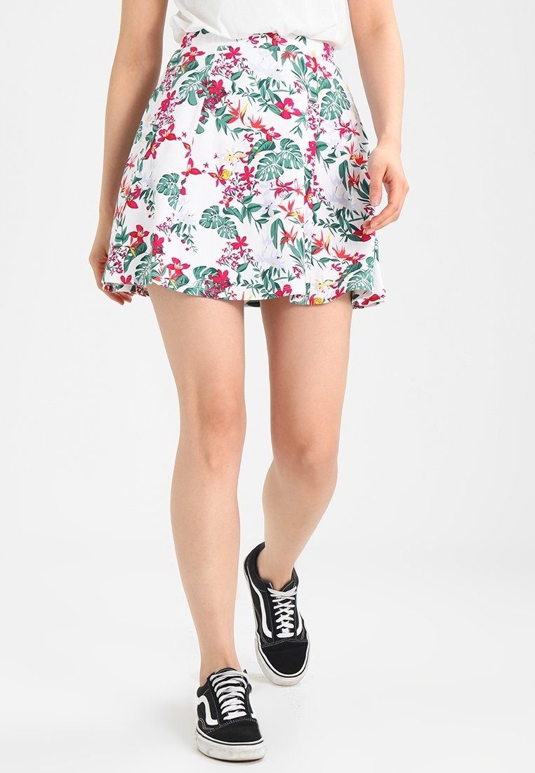 Femme portant une mini-jupe blanche à fleurs rouges, vertes et jaunes, associée à des baskets noires et blanches, debout devant un fond uni.