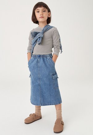 Enfant aux cheveux bruns courts portant une chemise rayée à manches longues, une jupe midi en denim, une veste en denim nouée autour des épaules, des chaussettes beiges et des sabots marron.