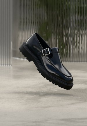 Scarpa nera in pelle lucida con punta tonda, cuciture decorative e cinturino con fibbia su suola spessa in gomma.