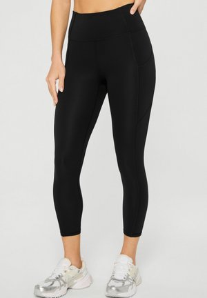 Woman wearing black high-waist cropped leggings with side pockets and white athletic shoes, standing against a plain background.