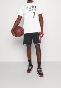 Camiseta blanca de Nike con "BKLYN NETS" y el número 7 impresos en negro. Pantalones cortos negros con rayas rojas a los lados. Sosteniendo un balón de baloncesto, lleva zapatillas negras.