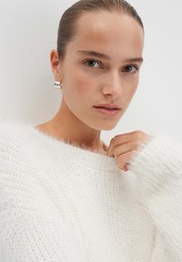 Witte gebreide sweater met een pluizige textuur. Model draagt zilveren ringoorbellen met een gladde, reflecterende oppervlakte.