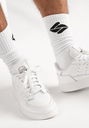 Witte sportieve schoenen met een glad leren bovenwerk, ronde neus en platte zool. Gecombineerd met witte geribbelde sokken met een zwart logo.