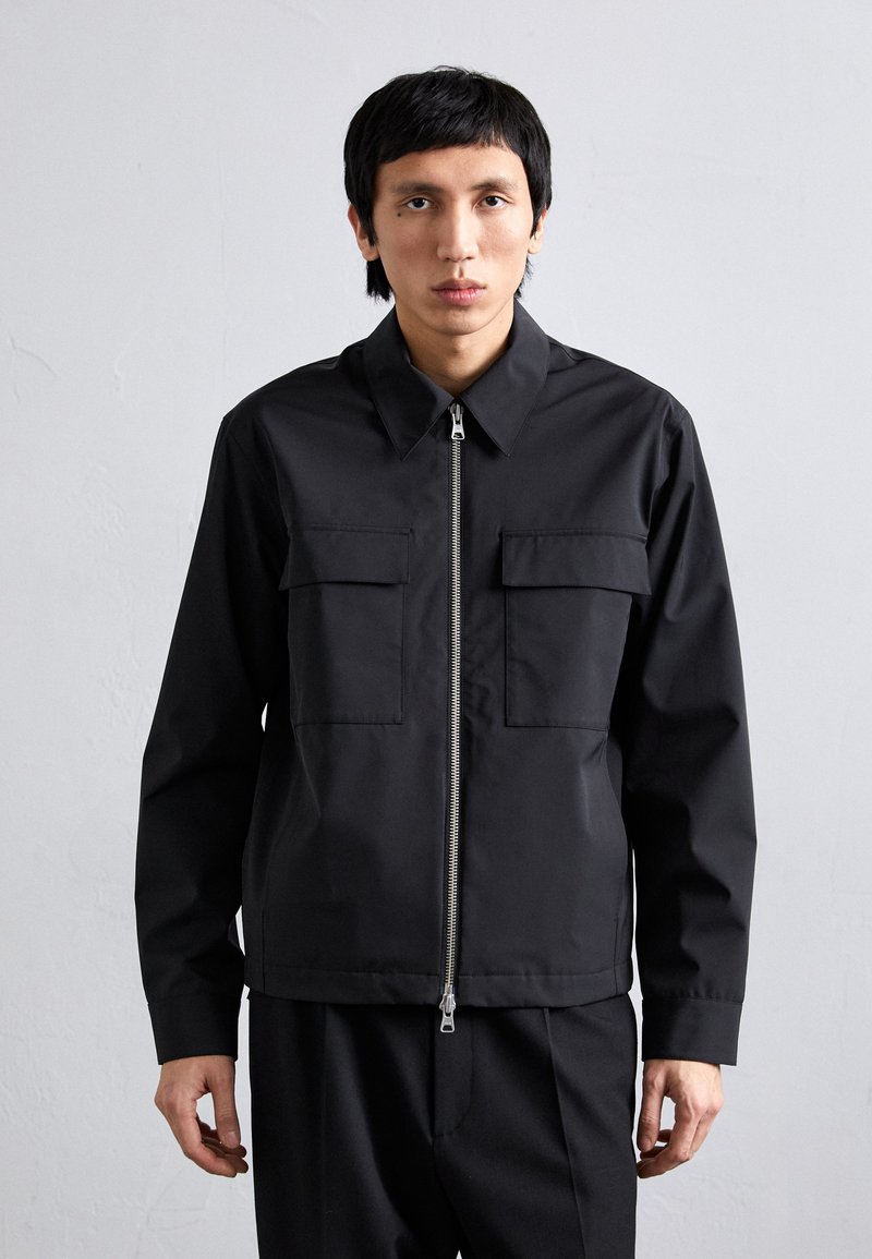 Svart zip-up jacka med krage, två bröstfickor och långärmar. Tillverkad av slät tyg med en strukturerad passform och minimalistisk design.