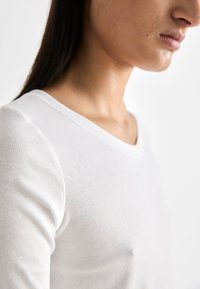 Top bianco a maniche lunghe con scollatura a V. Il tessuto sembra morbido e aderente, che mette in risalto una clavicola e un décolleté snelli.