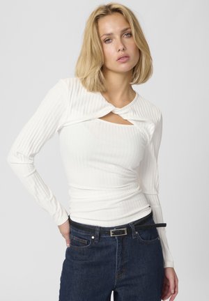 Weißes, geripptes Langarmoberteil mit einem Twist-Design am Kragen, das ein Cut-Out-Detail aufweist. Kombiniert mit dunkelblauen Jeans und einem schwarzen Gürtel.