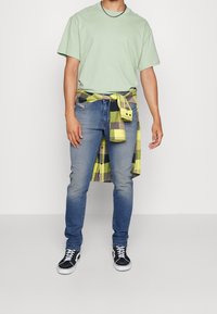 Světle zelené bavlněné tričko doplněné modrými džíny a žlutočernou kostkovanou košilí uvázanou kolem pasu. Černé tenisky dokončují celý outfit.