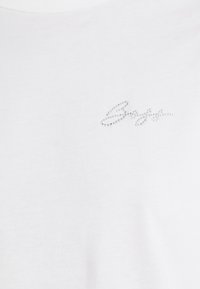 Camiseta blanca de algodón suave, con un logo en cursiva adornado con pequeños rhinestones en la parte delantera. Cuello redondeado.