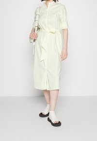 Vestito button-down a righe giallo chiaro con cintura, lunghezza fino al polpaccio. Abbinato a scarpe aperte con suola nera e tomaia bianca.