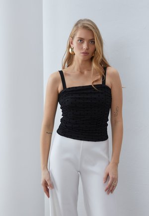 Camisola nera arricciata con texture a pois e spalline larghe, abbinata a pantaloni bianchi a vita alta. Orecchini a cerchio dorati completano il look.