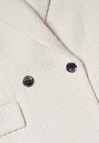 Hellbeige strukturierter Blazermaterial mit einem gewebten Muster; verfügt über zwei schwarze runde Knöpfe und einen Reverskragen.