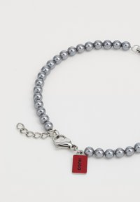 Grå pärlhalsband med silverlås och röd rektangulär tagg med texten "HUGO". Slät, glänsande yta genom hela smycket.
