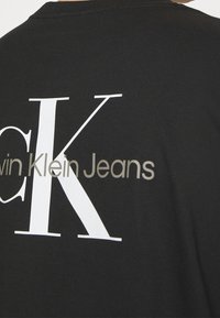 Černé tričko s velkým bílým nápisem "CK" a menším béžovým logem "Calvin Klein Jeans" na zádech.