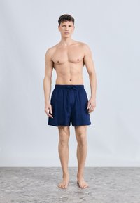 Μυώδης άντρας που στέκεται ξυπόλυτος σε πέτρινο δάπεδο, φορώντας σκούρο μπλε μαγιό, με φόντο απλό ανοιχτό γκρι.